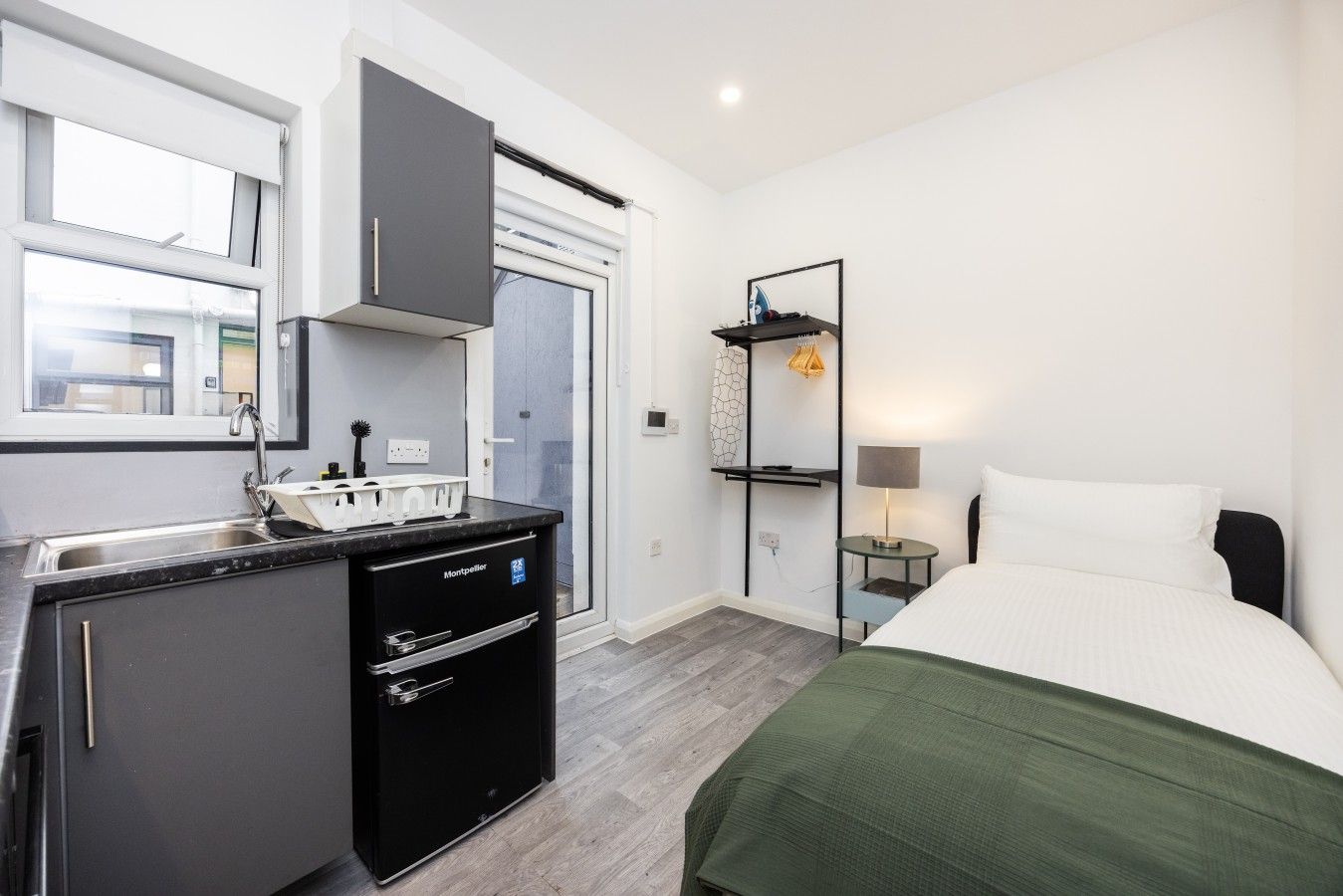 Una habitación de hotel compacta con kitchenette, una cama con manta verde, una mesita de noche con lámpara y un pequeño estante con adornos.