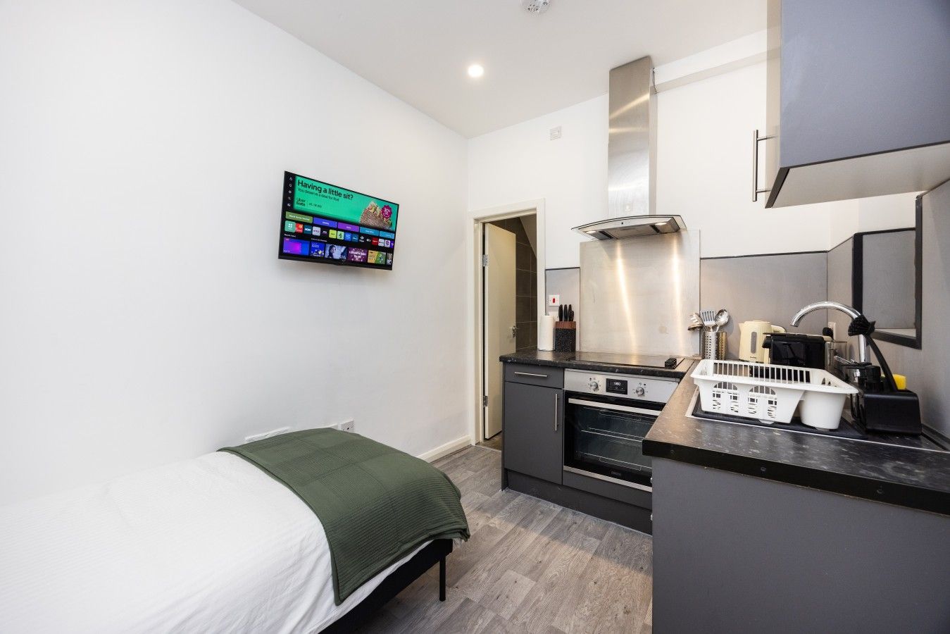 A compact room with a bed, a kitchenette, and a TV mounted on the wall.