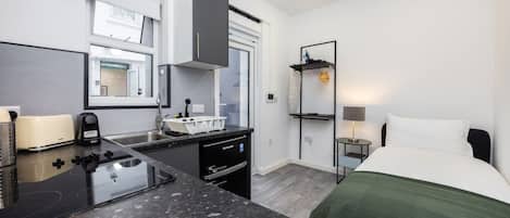 Habitación de hotel compacta con kitchenette, una cama con manta verde, una mesita de noche con lámpara y una ventana con vistas al exterior.
