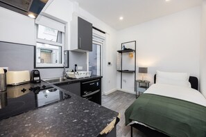A compact hotel room with a kitchenette, a bed with a green blanket, a bedside table with a lamp, and a window with a view of the outside. - Earl`s Court Residence | Elegant Studio | London (Greater London)