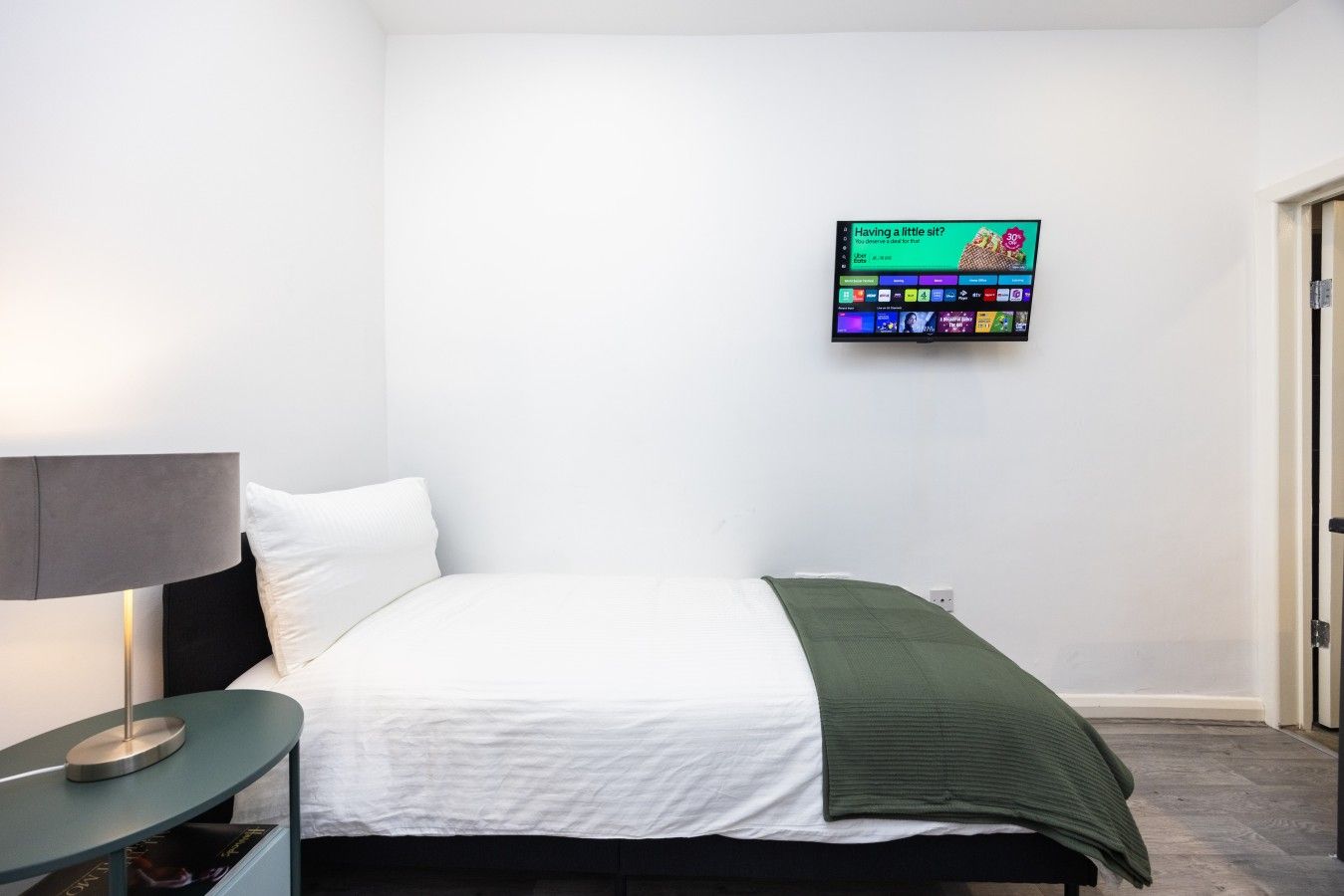 A hotel room with a bed, a nightstand, a lamp, and a TV mounted on the wall displaying a program.