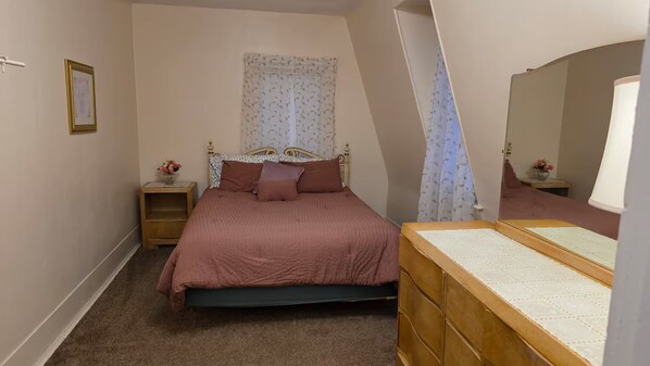 A bedroom with a bed, bedside table, dresser, mirror, and a window with floral curtains.