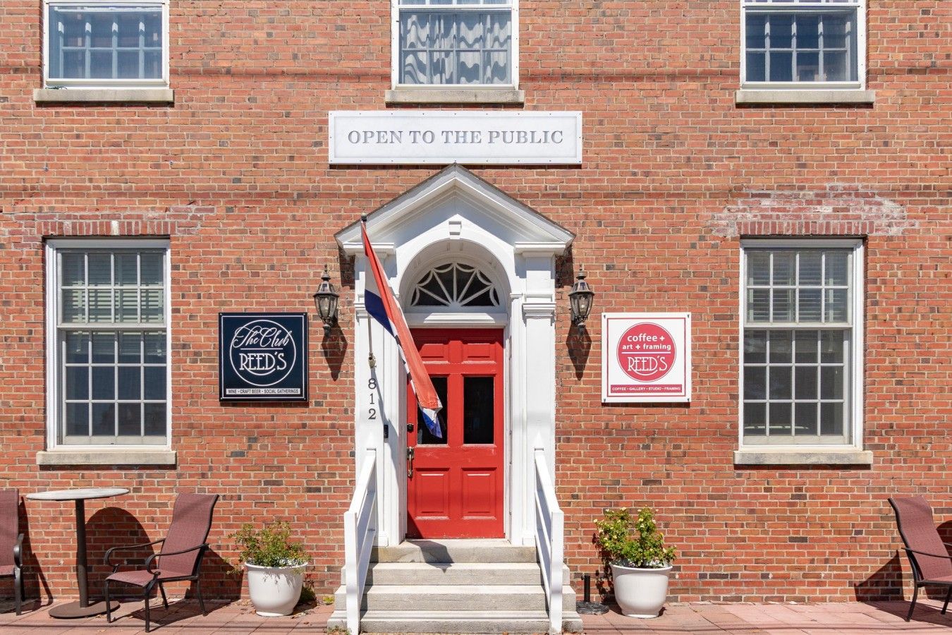 Un bâtiment en briques avec une porte rouge, une enseigne indiquant « OUVERT AU PUBLIC » et deux autres enseignes faisant la publicité du « Club REED’S » et proposant du « café + art + encadrement ».