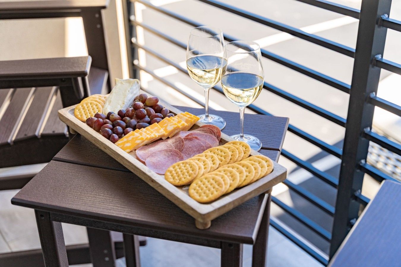 Una tabla de madera con queso, galletas y uvas sobre una mesa. También se ven dos copas de vino blanco.