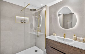 Ein modernes Badezimmer mit einer gläsernen Duschkabine, einem Waschtisch mit Holzschrank und einem großen Spiegel mit goldener Rahmung.