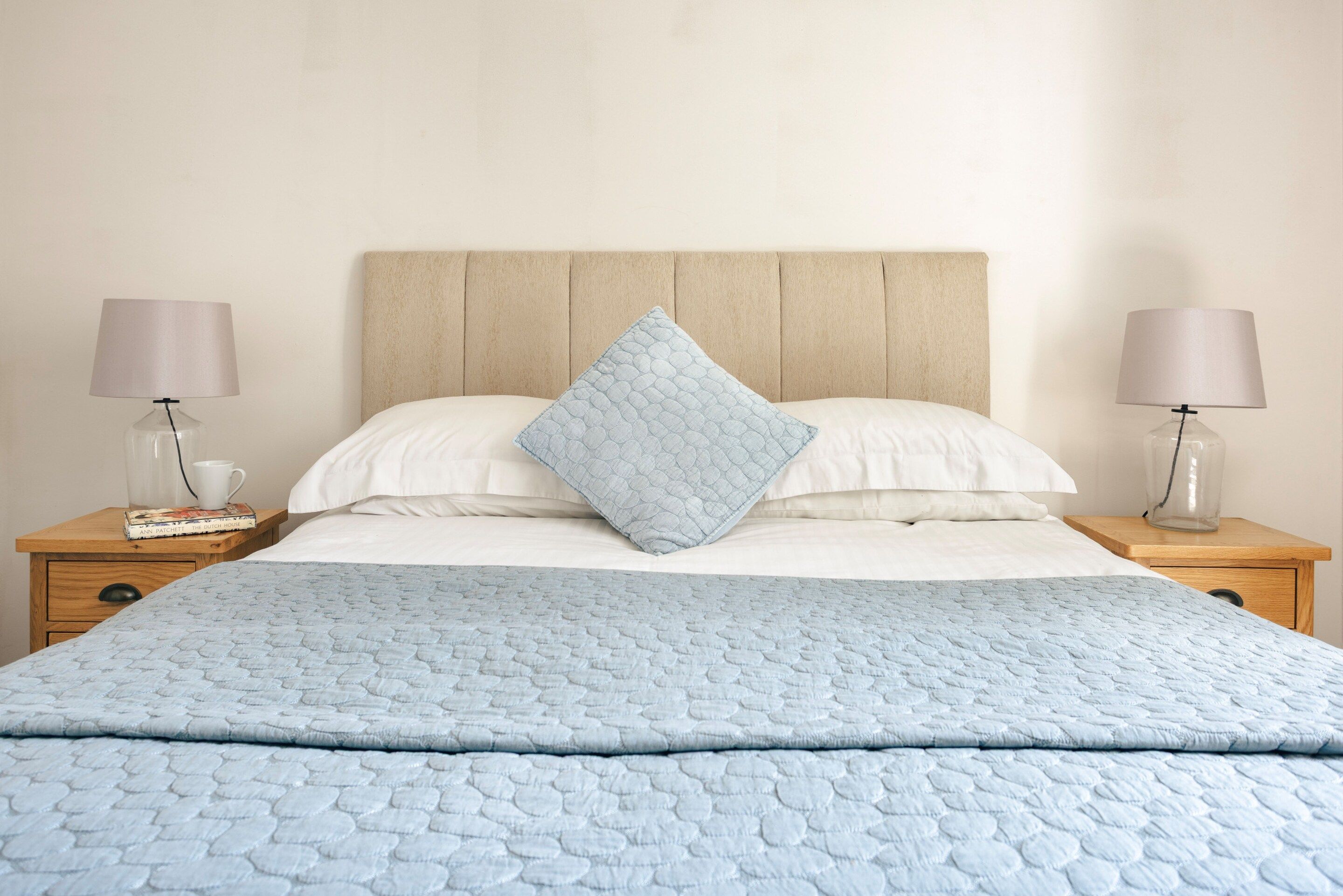 Een net opgemaakt bed met een blauwe quilt, twee bedlampen, een houten nachtkastje en een boek.