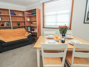 A living room with a sofa, a dining table with chairs, a bookshelf, and a window with blinds. - The Bookshop Apartment (Cowes)