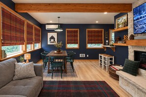 A cozy living room with a fireplace, a dining area with a wooden table and chairs, and large windows with plaid curtains.
