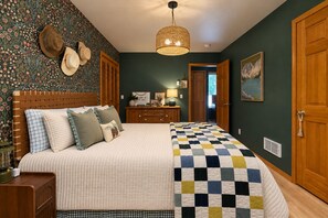 A bedroom with a large bed, a dresser, a nightstand, a wall-mounted lamp, and a door leading to another room.