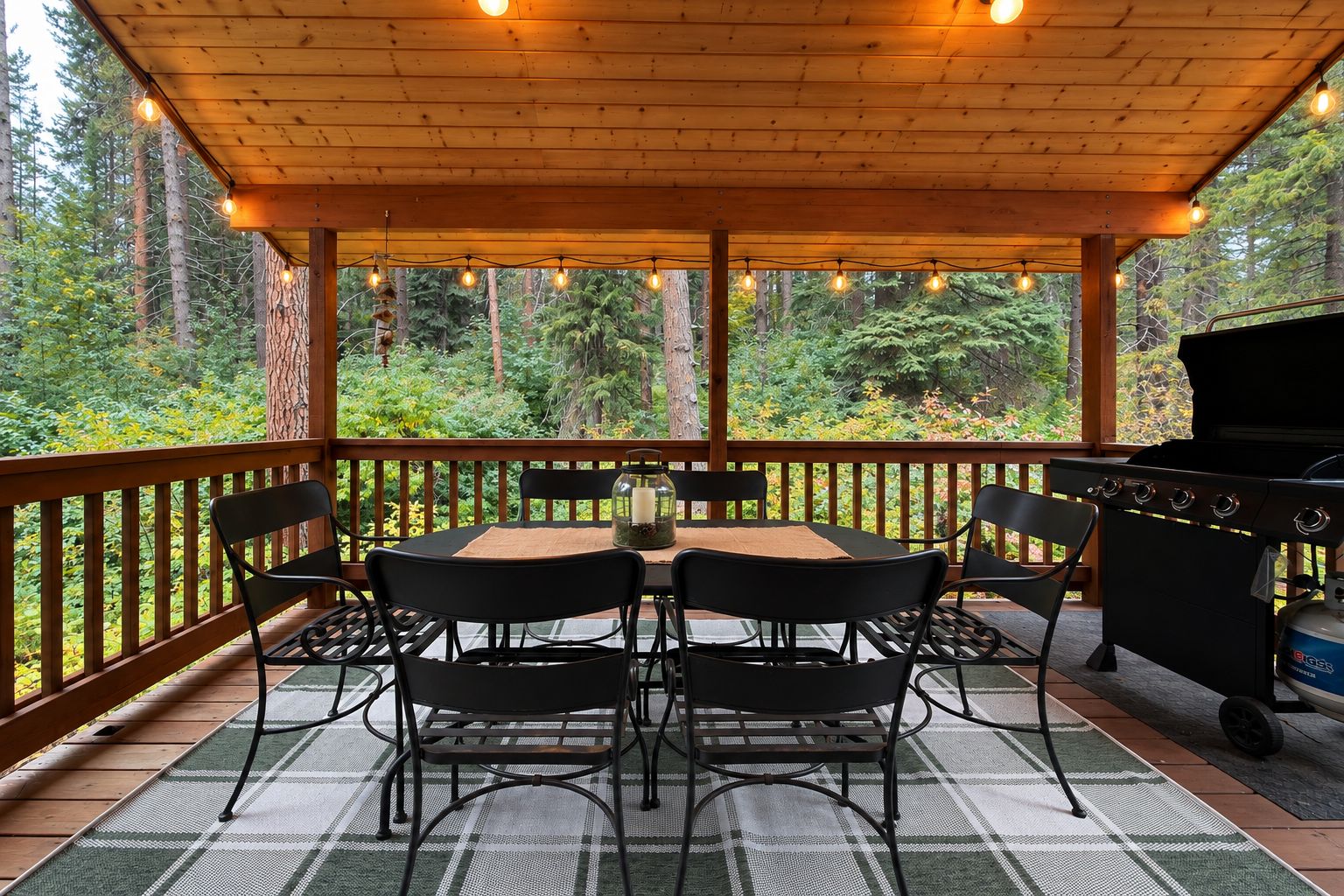 An outdoor patio with a round table and chairs, a grill, and a view of a forest.