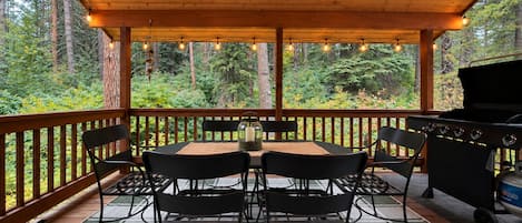 An outdoor patio with a round table and chairs, a grill, and a view of a forest.