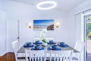 Un coin repas avec une table ronde, des chaises blanches, de la vaisselle bleue, un tableau sur le thème de la plage et une orchidée blanche.