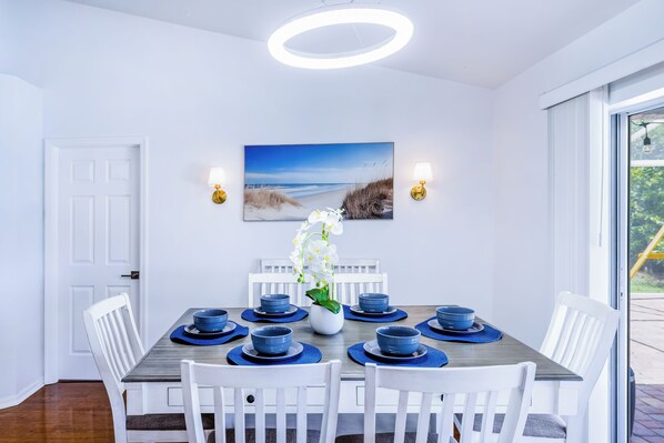 Et spiseområde med et rundt bord, hvide stole, blåt service, et maleri med strandmotiv og en hvid orkidé.