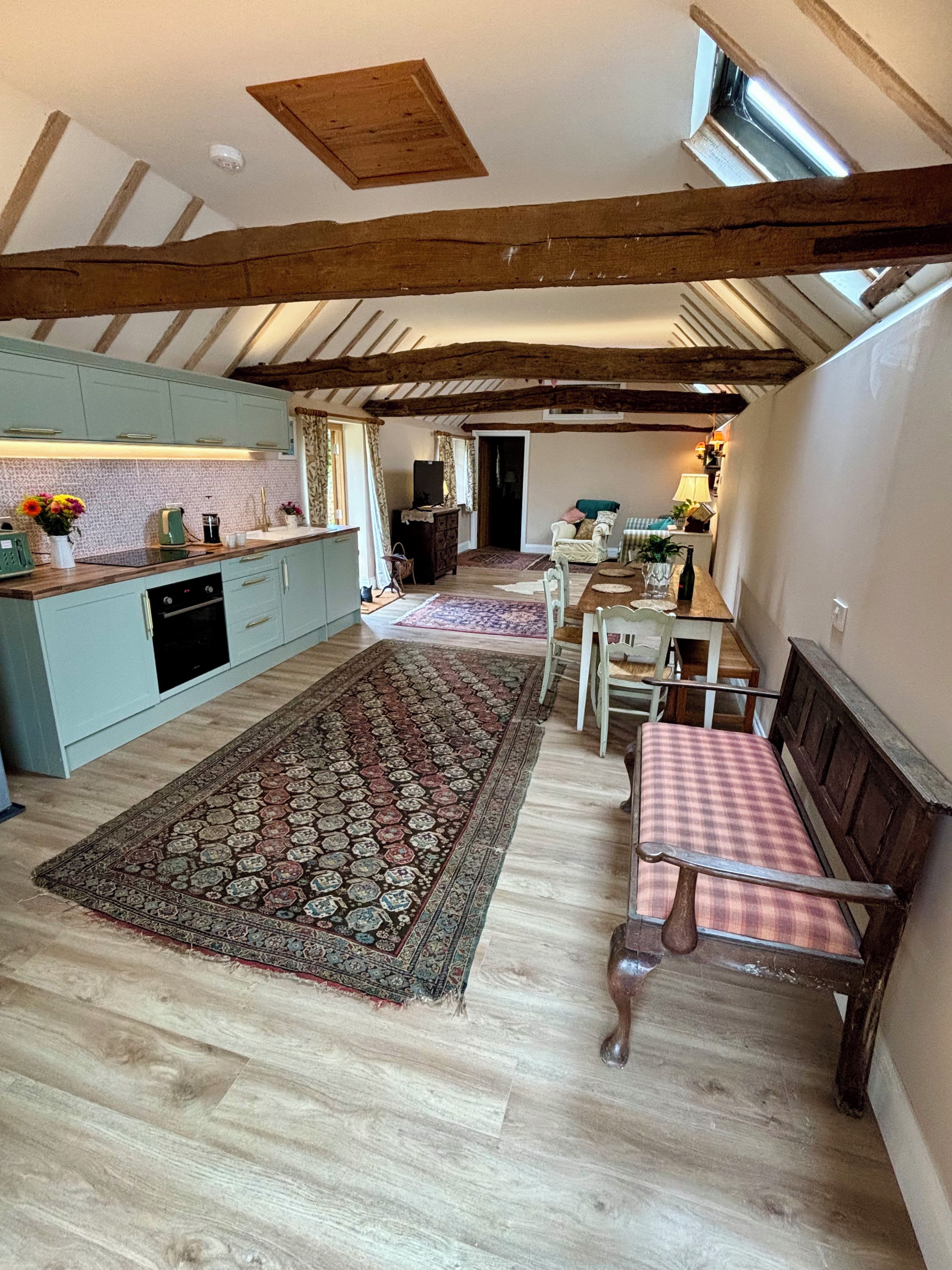 A spacious loft with exposed wooden beams, a kitchen with light blue cabinets, a dining area with a checkered table, and a large patterned rug.