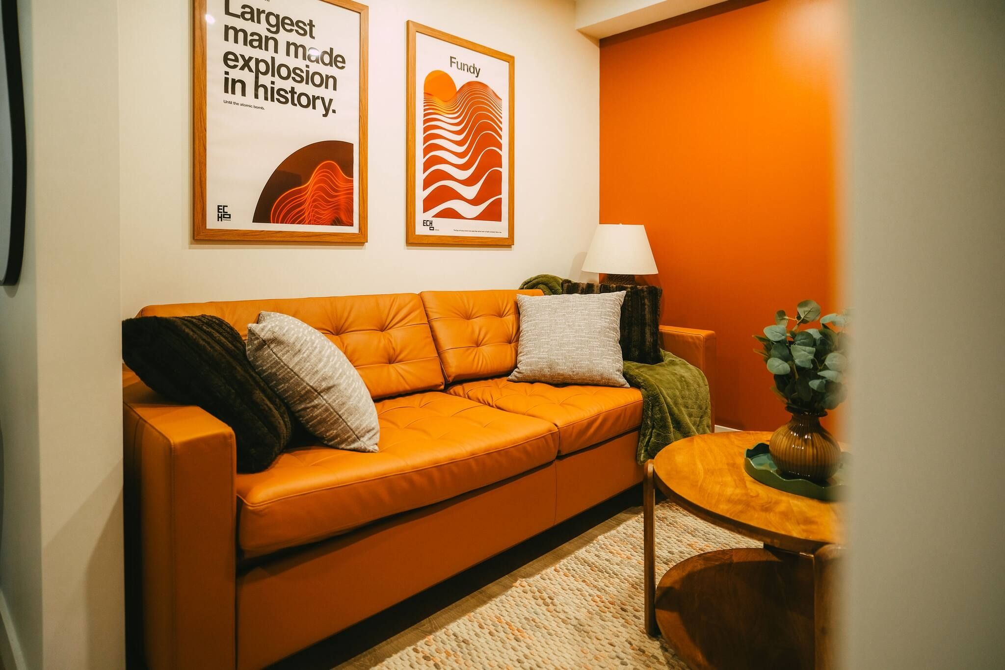 A room with an orange sofa, two framed artworks, a lamp, and a small table with a plant.