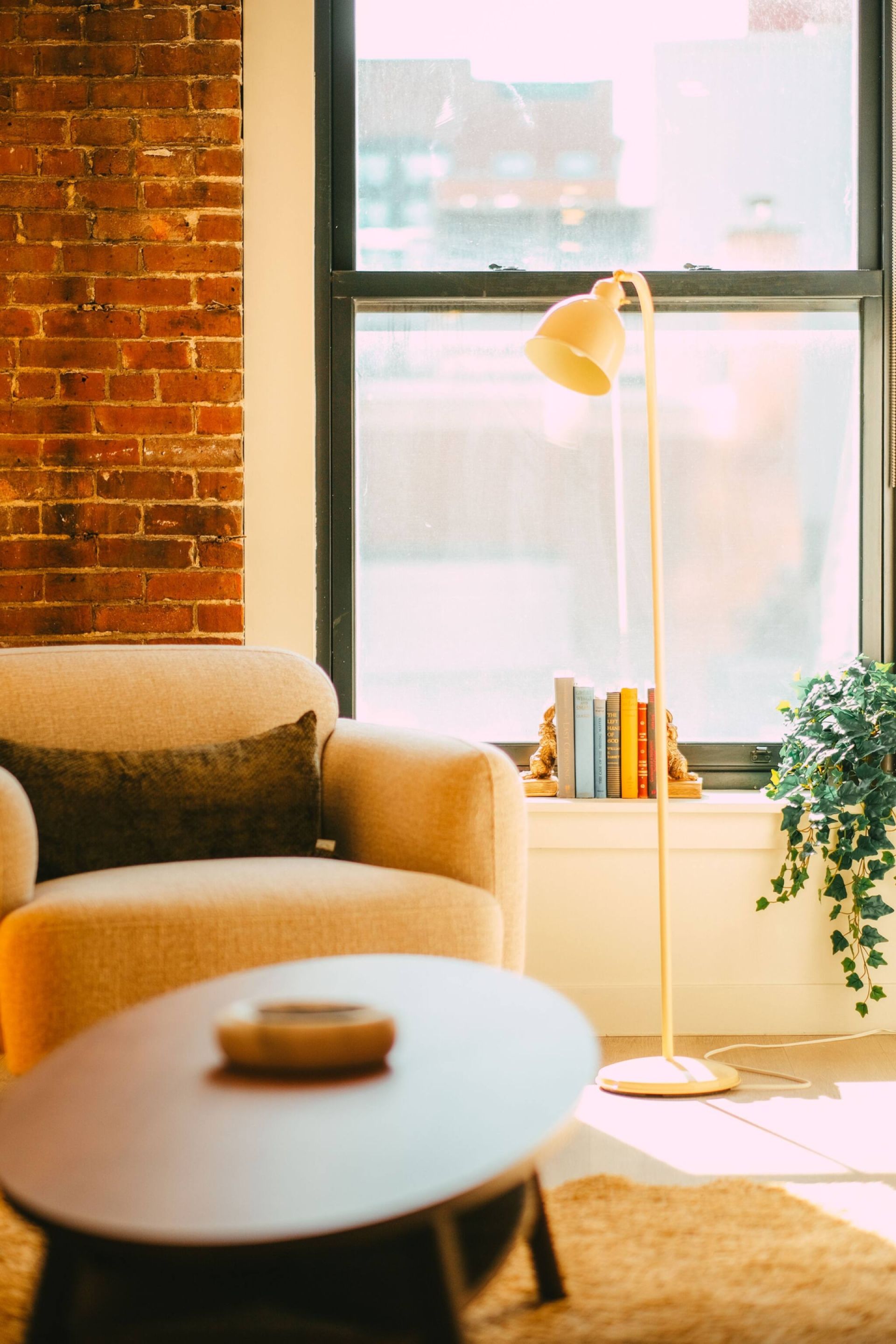 A cozy room with a brick wall, a window with a view, a floor lamp, a sofa, a coffee table, and a plant.