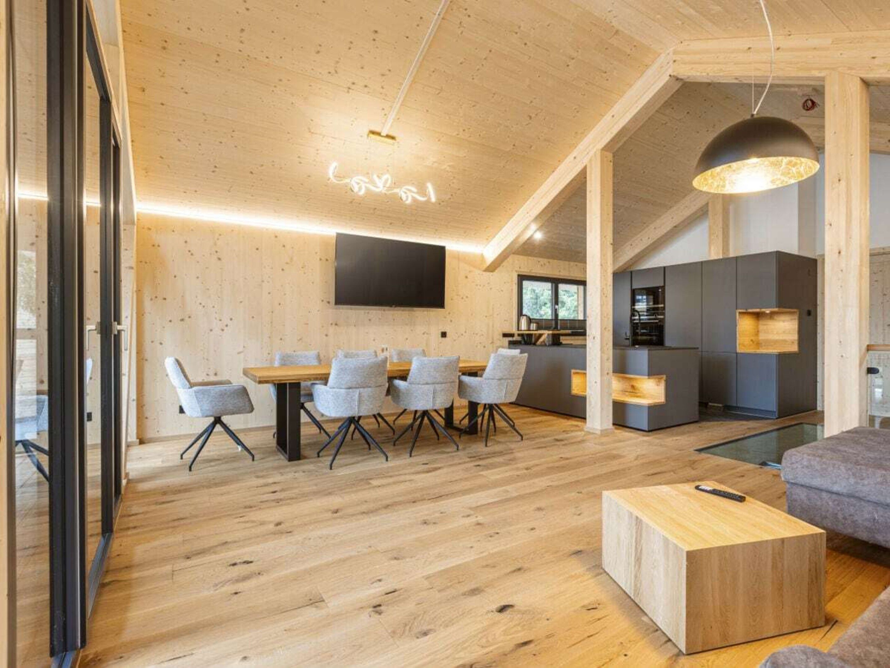 Un interior modern amb sostre de fusta, zona de menjador amb cadires grises, cuina amb armaris grises i un sofà gris.