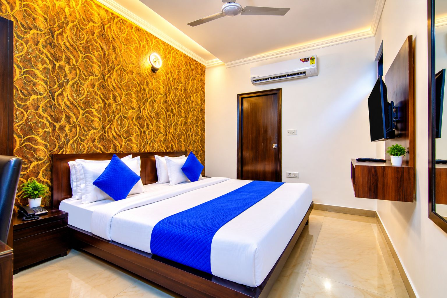 A hotel room with a bed, a ceiling fan, an air conditioning unit, a TV, and a wooden door.
