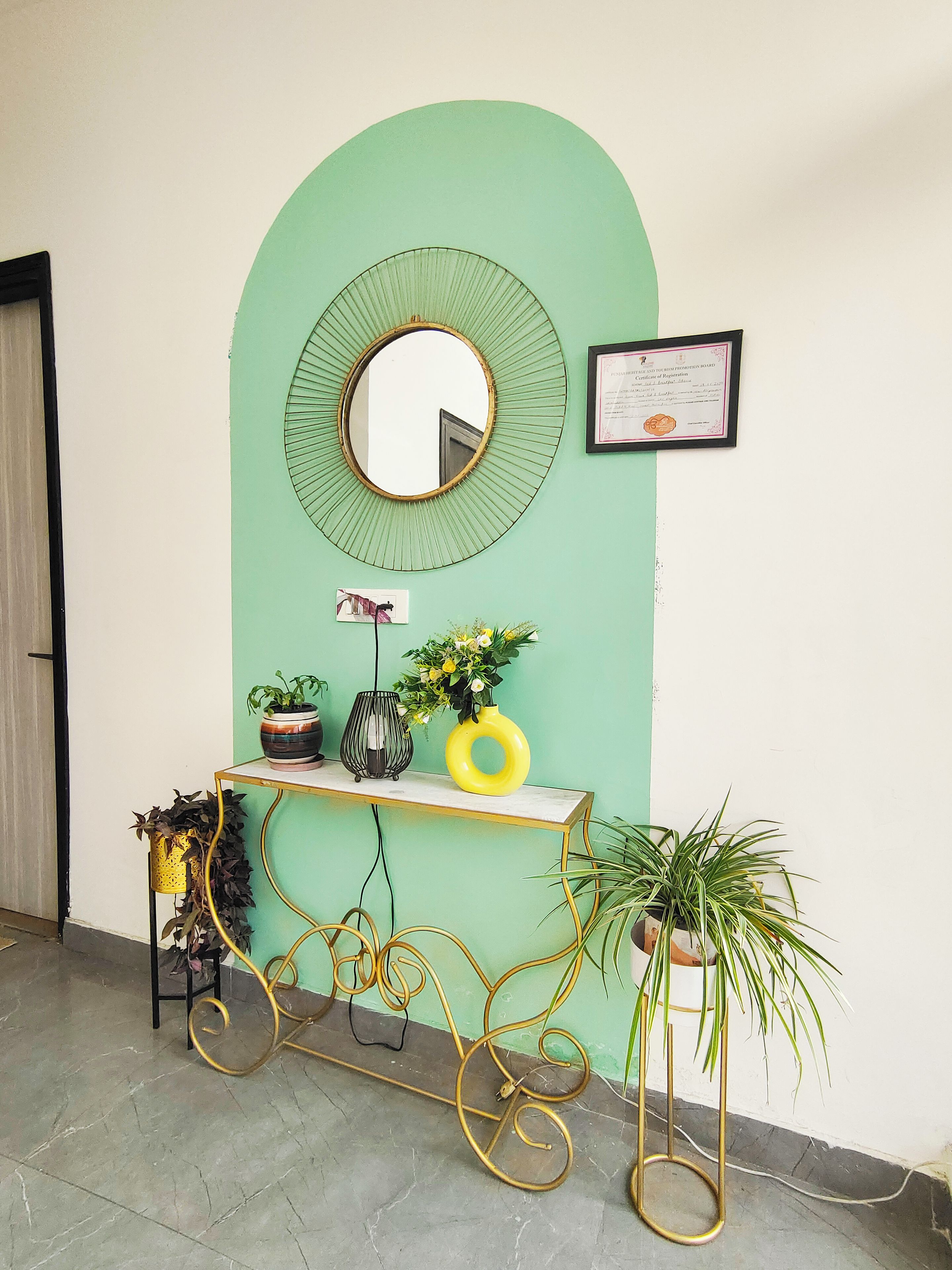 Una habitación con pared verde, un espejo redondo, un certificado enmarcado, una pequeña mesa con plantas en macetas y una silla dorada.