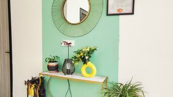 Una habitación con pared verde, un espejo redondo, un certificado enmarcado, una pequeña mesa con plantas en macetas y una silla dorada.
