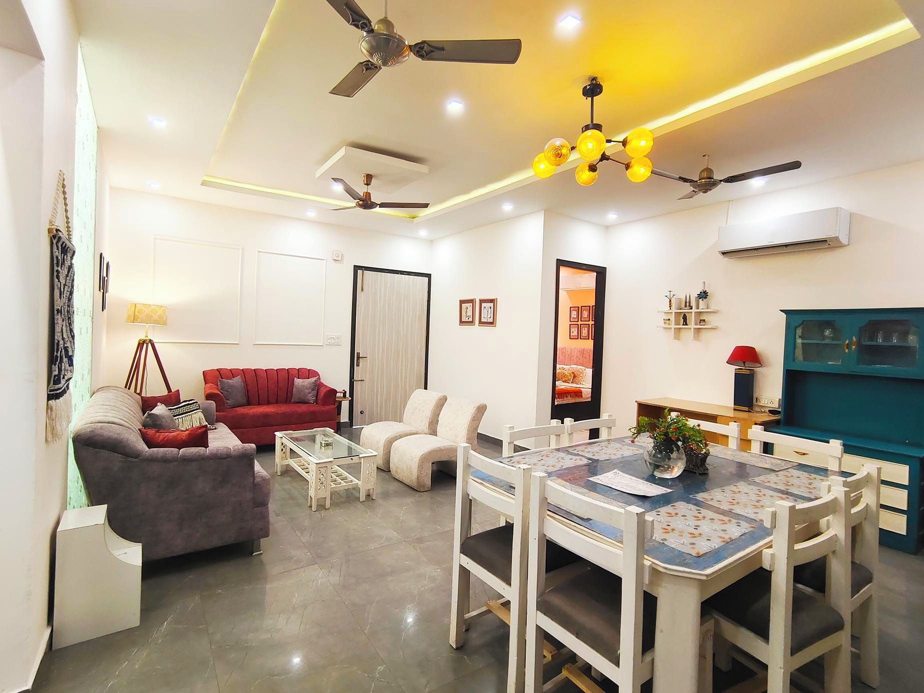 A modern living room with a red sofa, white armchairs, a glass coffee table, and a dining area with a blue table and white chairs.
