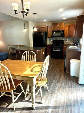 A dining area with a wooden table and chairs, a kitchen with wooden cabinets and appliances, and a white trash bin. - Raptor Retreat, 3BR/3Bath North Spokane Home Base (Spokane)