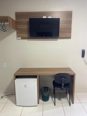 A compact room with a wooden desk, a small white refrigerator, a chair, and a mounted television.