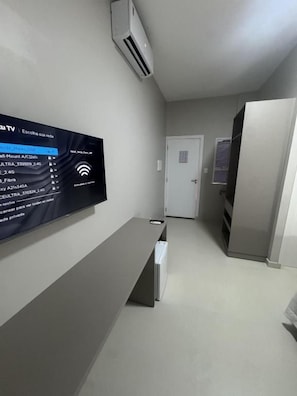 A modern hotel room with a flat-screen TV mounted on the wall, a built-in air conditioning unit, a door, and a refrigerator.
