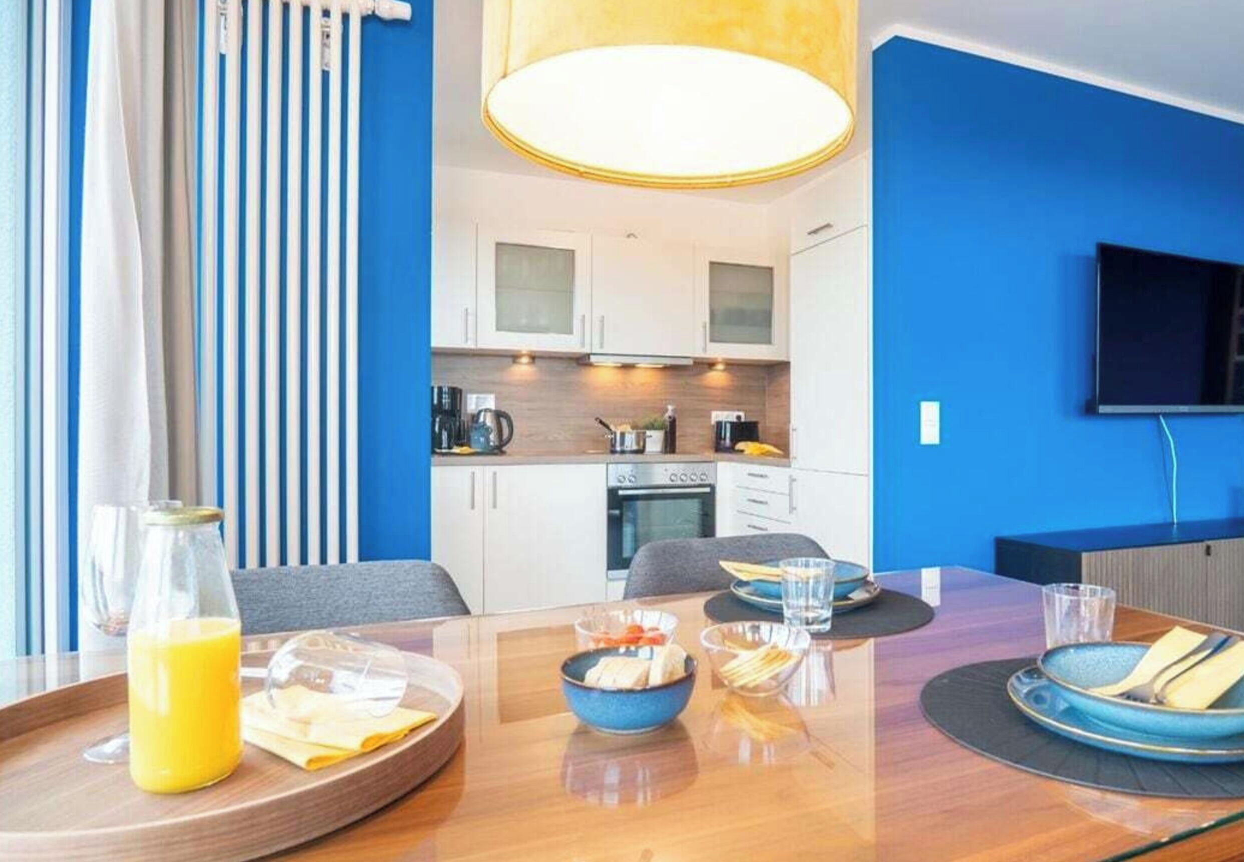 A modern hotel room with a dining table set for breakfast, a kitchen area, and a TV mounted on the wall.