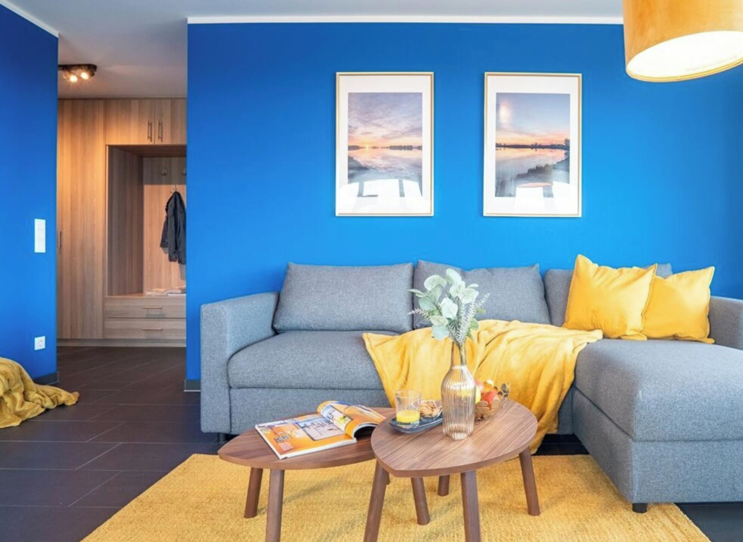 A living room with a blue wall, a grey sofa, yellow cushions, a coffee table with a magazine, and two framed pictures.