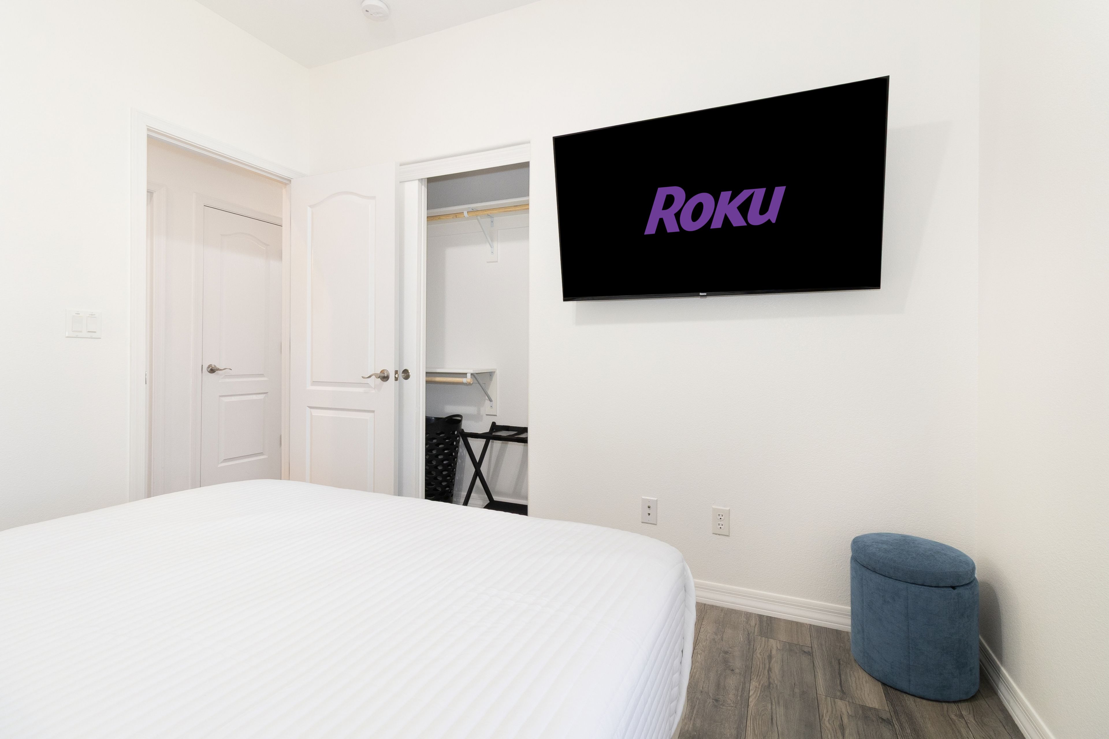 A bedroom with a bed, a TV mounted on the wall displaying 'Roku', a closet, and a small blue ottoman.