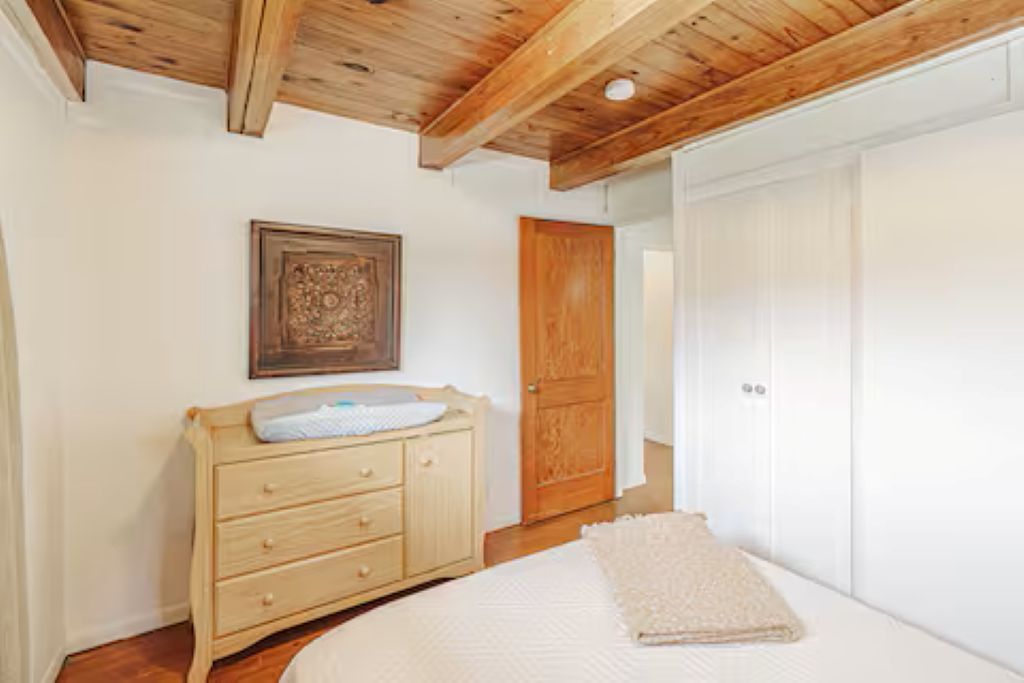 A bedroom with a wooden ceiling, a wooden dresser, a bed with a white blanket, and a white wardrobe.