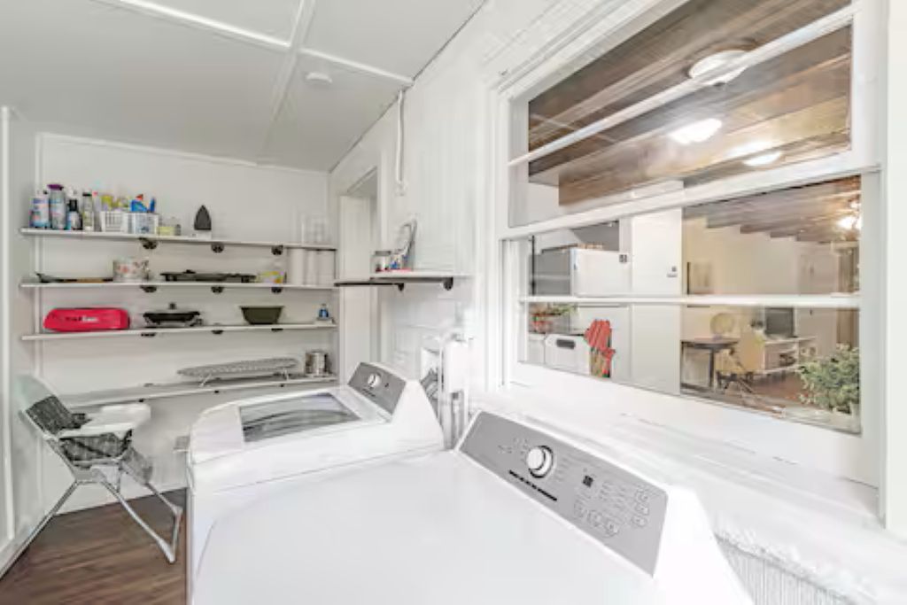 Een wasruimte met een wasmachine, planken met schoonmaakspullen en een raam met uitzicht op het interieur.