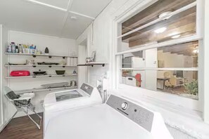Una zona de bugaderia amb una rentadora, prestatges amb productes de neteja i una finestra que dona a l'interior.