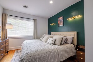 A bedroom with a bed, a dresser, a window with curtains, a wall-mounted light fixture, and a painting. - Stylish 2BR apt in beautiful Petersburg w/ parking, laundry, & outdoor space (Petersburg)