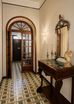 Un pasillo con piso estampado, una puerta de madera con paneles de vidrio, un espejo fijado a la pared y una mesa con una escultura de busto.