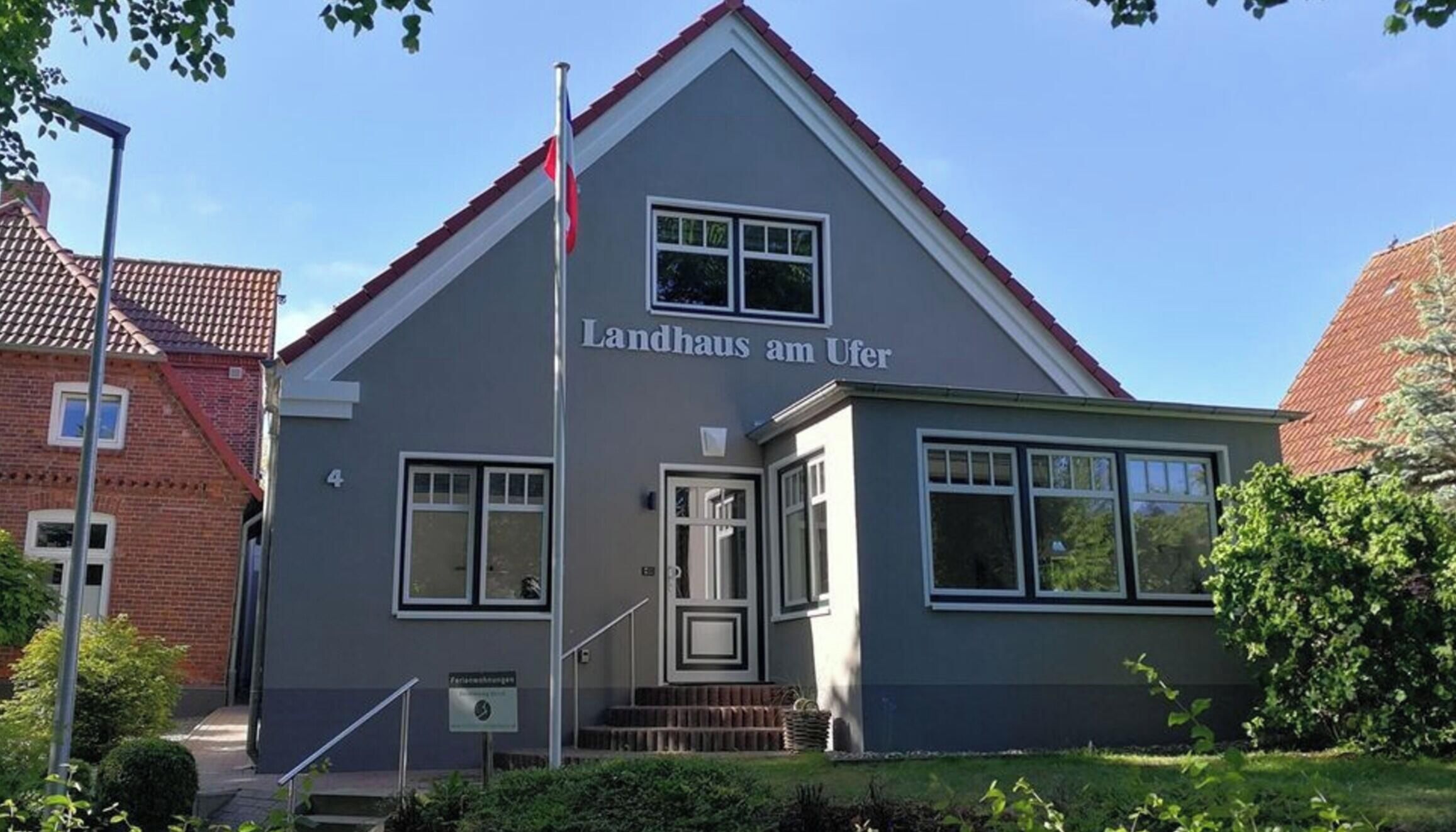 A building with a grey facade, a red roof, and a sign that reads 'Landhaus am Ufer'.