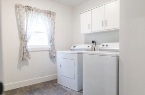 A laundry room with two white washing machines, a window with patterned curtains, and a cabinet above the machines.