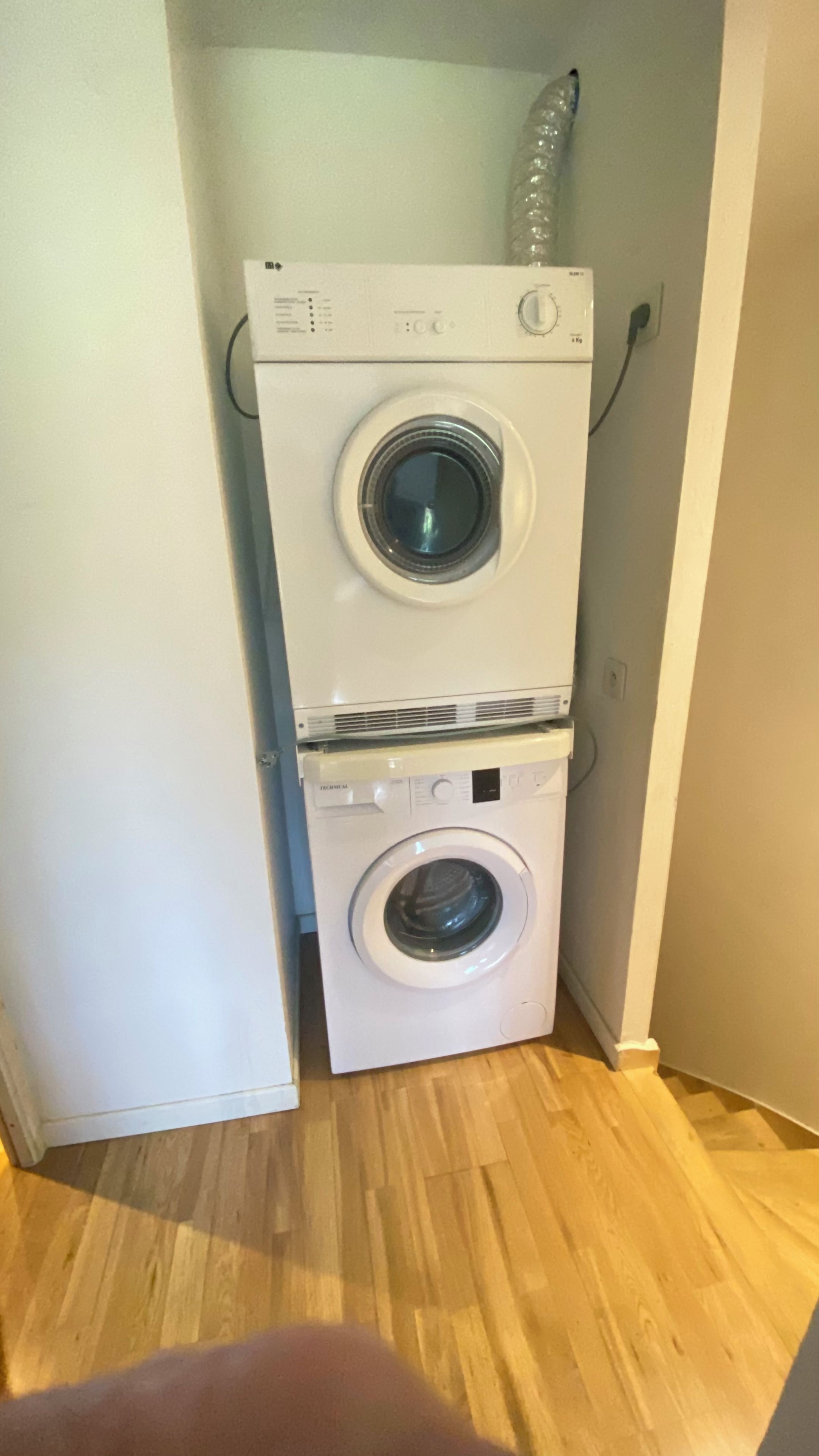 A stacked washer and dryer unit in a corner of a room with wooden flooring.