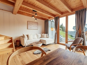 A cozy living room with a wooden ceiling, a white sofa, a wooden dining table, and a view of the outdoors through large windows. - Cabin in Niederwolz With Sauna (Oberwölz)