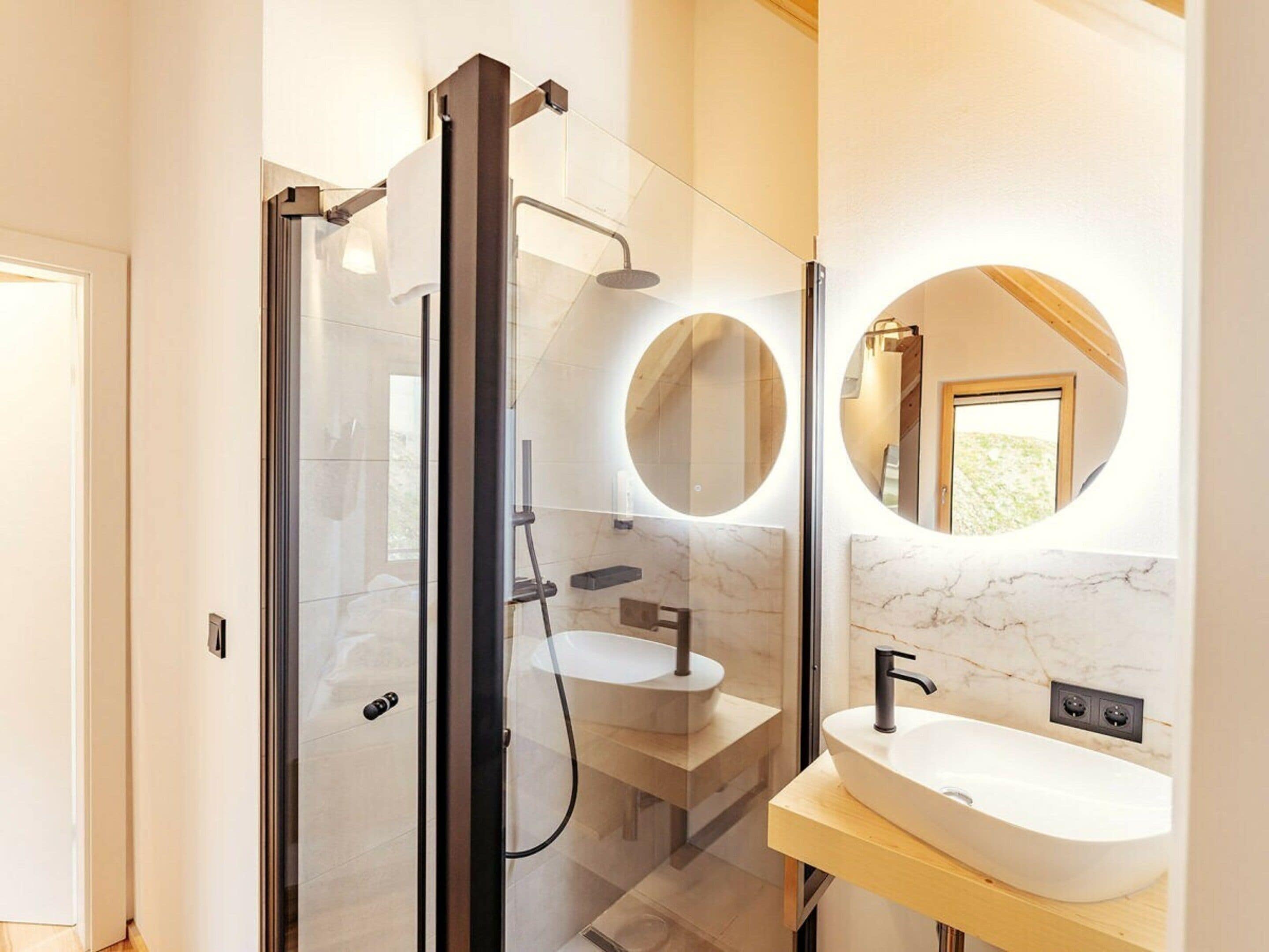 Un baño moderno con ducha de vidrio, espejo redondo y pared con azulejos de mármol.