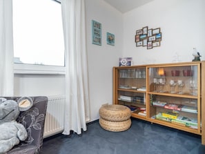 A room with a sofa, a bookshelf, a window with curtains, and a wall with framed pictures. - Ohana Holiday Apartment (Zingst)