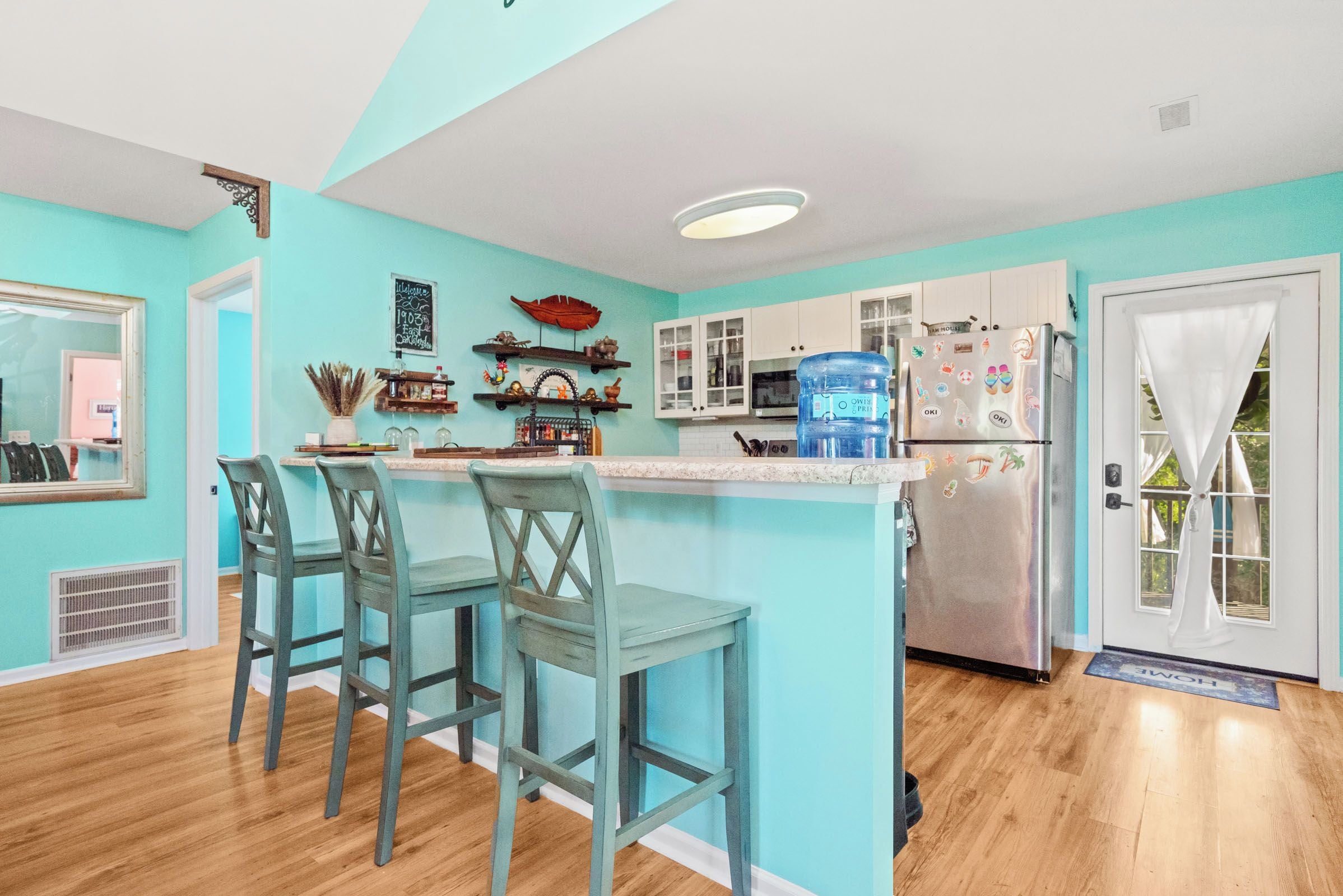 A kitchen with teal walls, wooden bar stools, a large water dispenser, and a stainless steel refrigerator.