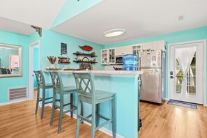 Une cuisine aux murs de couleur turquoise, avec des tabourets de bar en bois, un grand distributeur d’eau et un réfrigérateur en acier inoxydable.