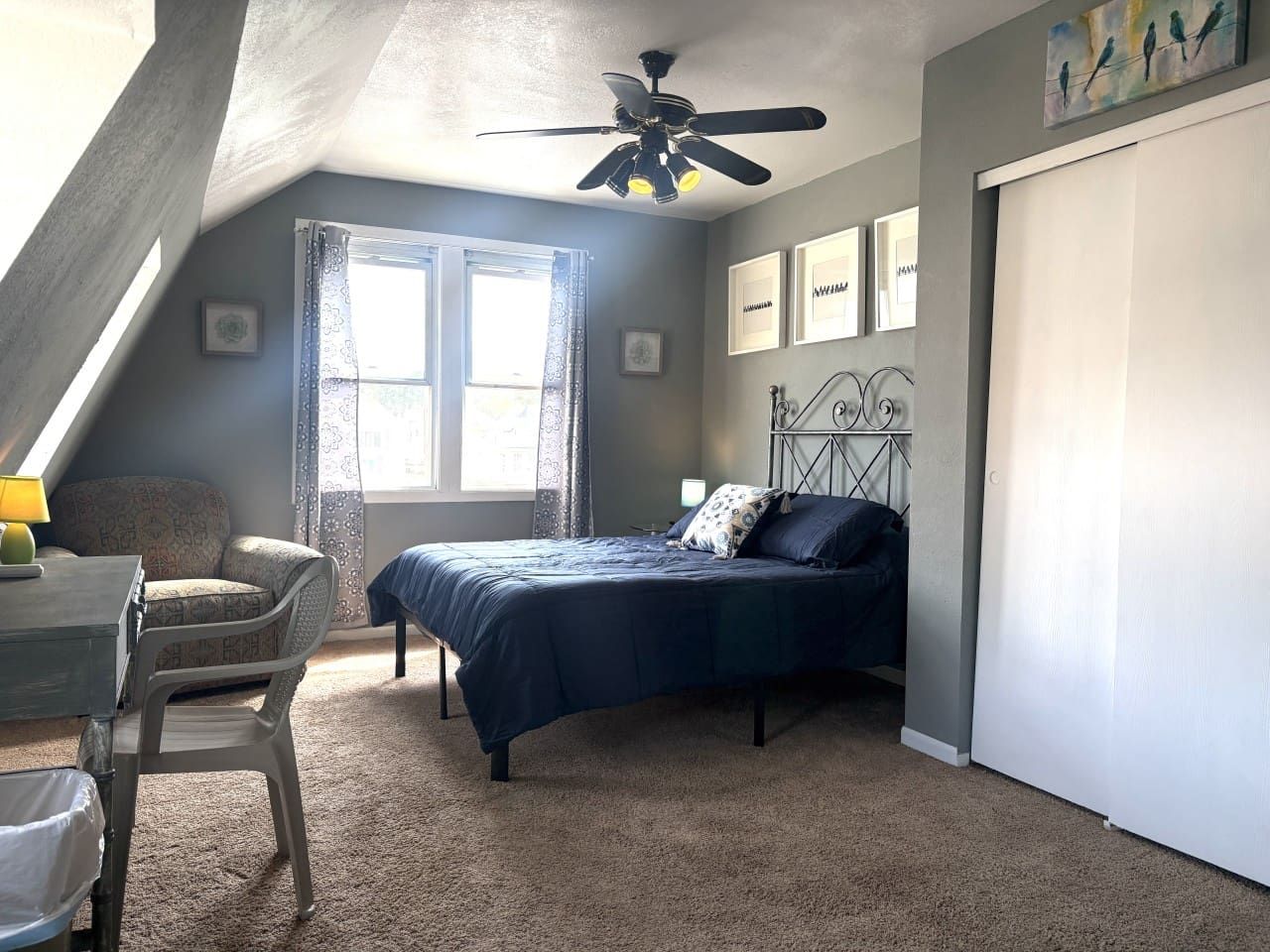 A bedroom with a bed, a desk, a chair, a ceiling fan, and a window with curtains.