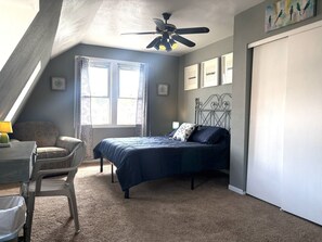 Une chambre à coucher comprenant un lit, un bureau, une chaise, un ventilateur de plafond et une fenêtre avec des rideaux.