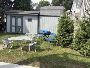 Eine kleine Gartenhütte mit Tür und Fenstern, ein runder Metalltisch mit Stühlen und ein Grill.