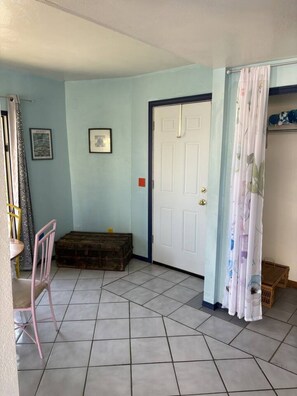 Ein Zimmer mit Fliesenboden, einer weißen Tür, einer kleinen Holzkiste, einem pinken Stuhl und einem Vorhang mit Blumenmuster.