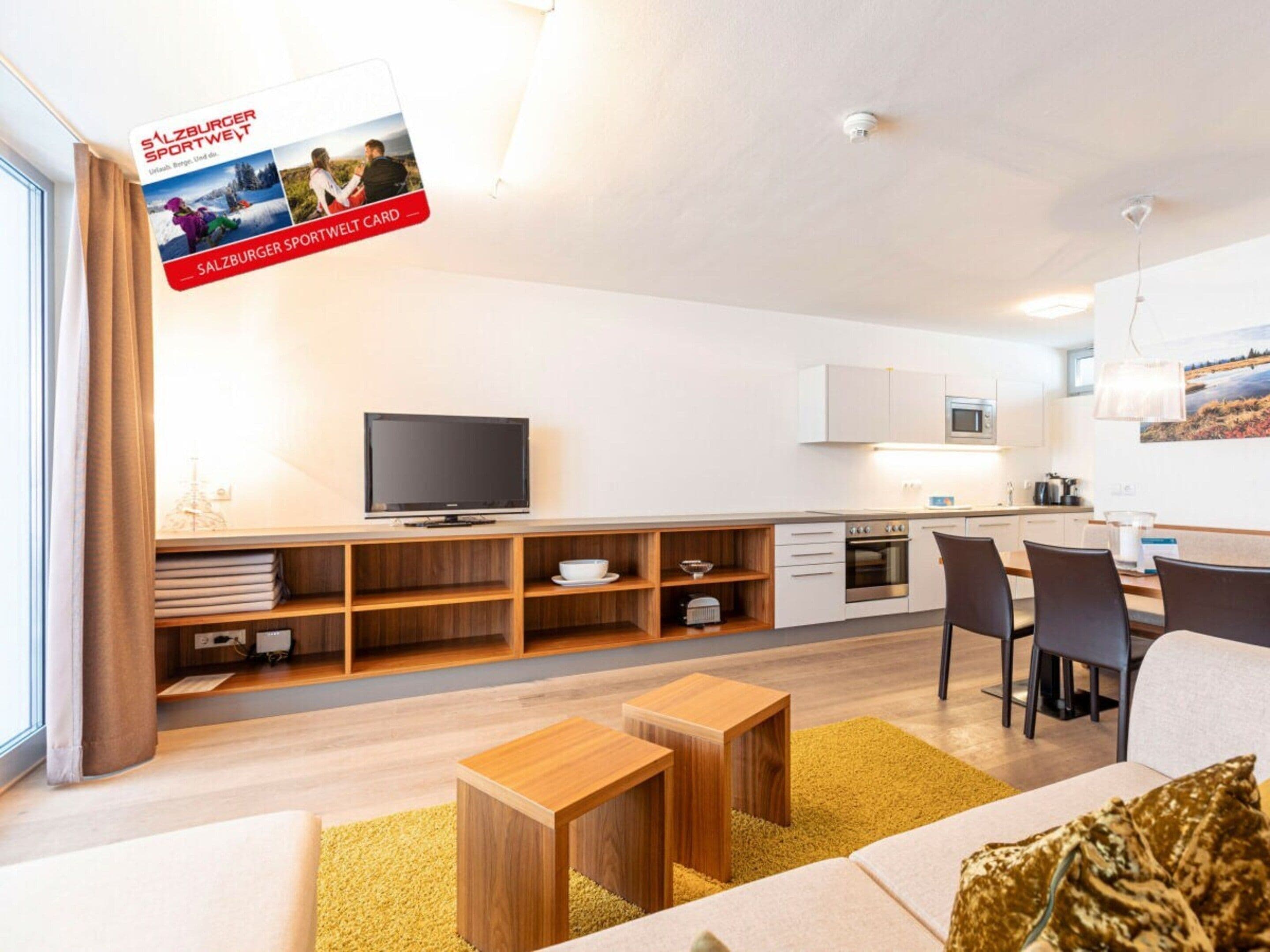 A modern living room with a television, wooden furniture, and a kitchen area in the background.