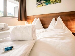 Ein Hotelzimmer mit einem ordentlich bezogenen Bett, einem aufgerollten weißen Handtuch und einem blauen Gegenstand auf dem Bett.